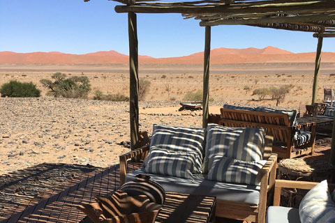 Communal seating area on a decking at Kulala Desert Lodge with comfy looking cushioned seats looking out over a panoramic landscape view the Namib desert and onto the red sunes of Sossuvlei in the background.