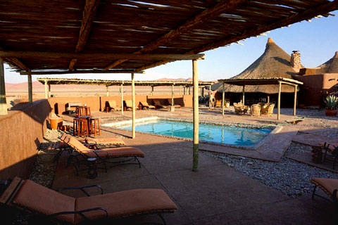 Swimming pool area at Kulala Desert Lodge Sossuvlei Namibia covered by a timeber and bamboo shade with seating and loungers arranged around the small pool in the centre and views out over the namib desert.