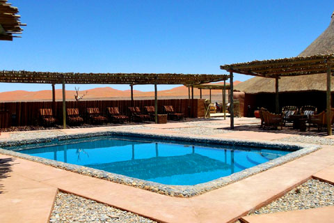 Swimming pool area at Kulala Desert Lodge Sossuvlei Namibia covered by a timeber and bamboo shade with seating and loungers arranged around the small pool in the centre and views out over the namib desert.