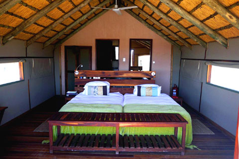 Inside the Kulala Desert Lodge chalets showing canvas walls and a comfy bed under a ceiling fan hanging from the centre of a high thatch roof. At the back behind the bed are doors leading to a wardrobe and a bathroom, and a full length mirrir. Down the sides the canvas blinds are rolled up allowing fresh air and light to shine in through mesh windows. 