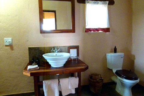 Bathroom in a Kulala Desert Lodge Chalet showing brick wall at the back with an open window above the fully plumbed toilet on the right hands side which has a wooden seat. On the left hand side is a porcelain sink with hot and cold taps above fixed onto a wooden stand which has fluffy white towels hanging down and a large mirror mounted above.