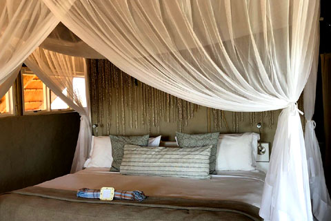 A large kingsize bed inside a guest villa at Little Kulala Camp. The bed is surrounded by mosquito nets hung from a square frame above and at the back of the photo you can see through the windows into the shaded thatch area beyond.