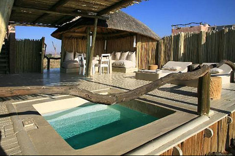 Private plunge pool area on the side verandah of a Little Kulala Camp guest villa. To the right of the plunge pool are two loungers, and at the back of the photo is a private sitting area with cusioned seating around the outside as well as a table with chairs. The plunge pool is covered by a bamboo shade, and to the right where there is no shade you can see the bright blue cloudless sky of Namibia.