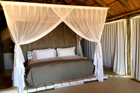 Inside the Little Kulala Camp guest villas showing a comfy 4 poster bed with mosquito nets under a igh thatch roof. Thick curtains cover the large windows down the side to help keep the guest villa cool. 