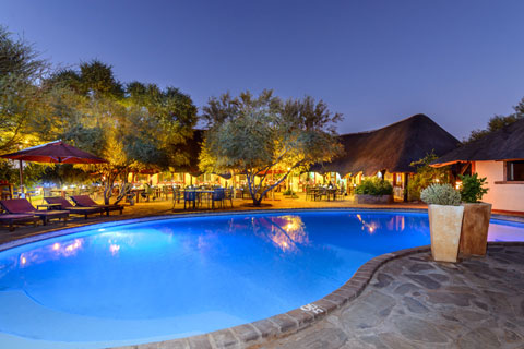 The sparkling swimming pool area at Namib Desert Lodge lit up at night byt the restaruant area behind showing tables and chairs set for diners and sun loungers by the cold water swimming pool.