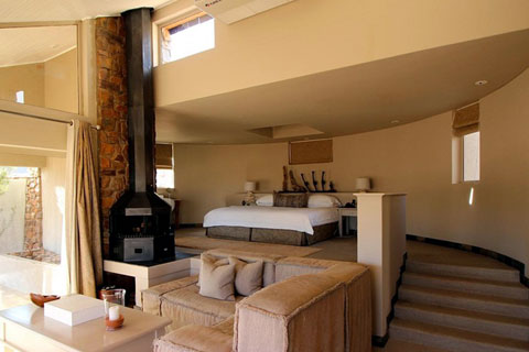 Inside view of one of the guest villas at Sossusvlei Desert Lodge. Looking up to the bedroom area from the living area which is open plan through to the bedroom which is on a higher level. There is an outside verandah on the left hand side and a short stairway on the right leading to a large king size bed on the next level.